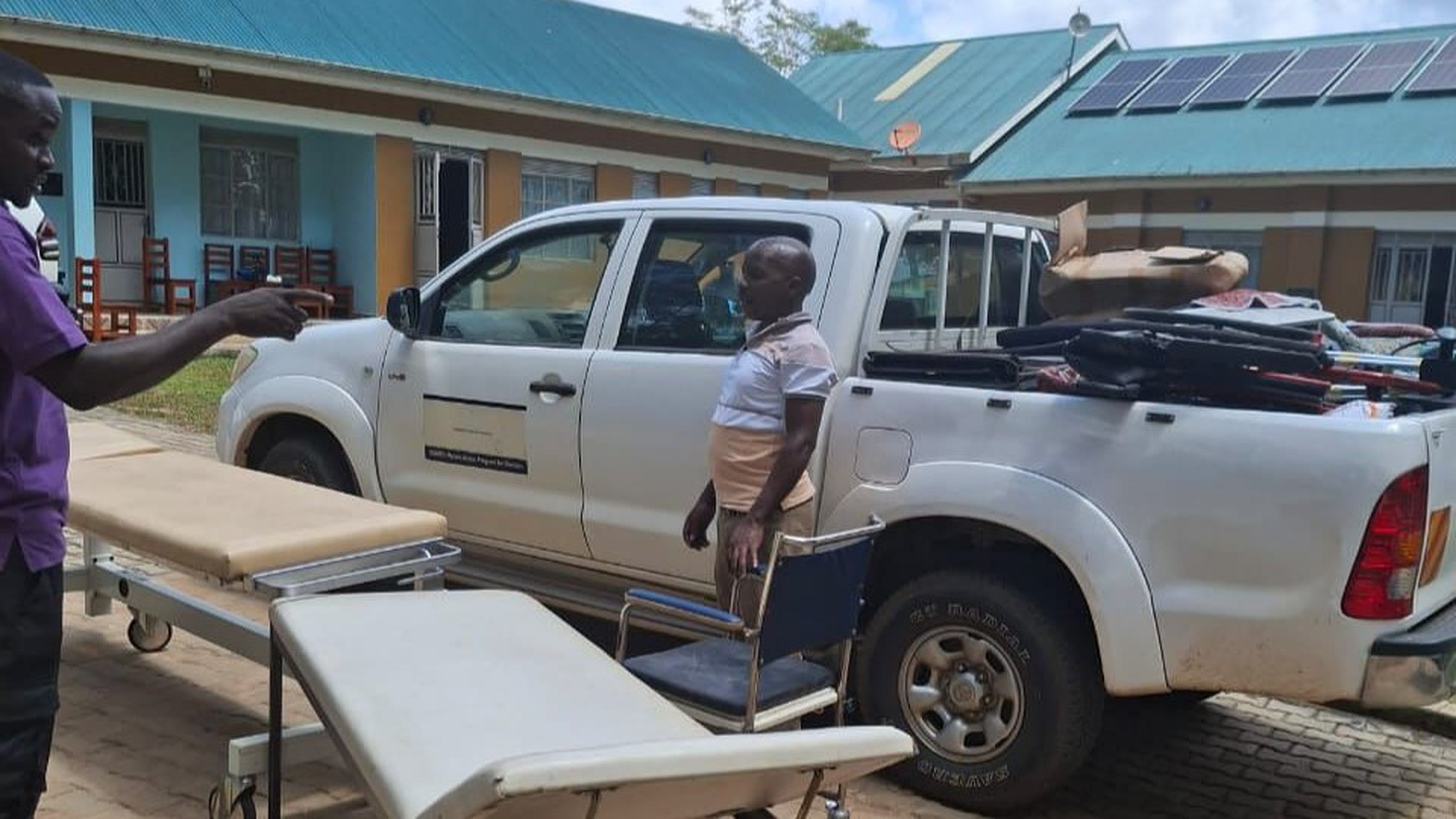 Ein mit Matratzen und Decken beladener Pick-up vor einer Gesundheitsstation in Uganda; davor stehen Untersuchungsliegen und ein Rollstuhl.