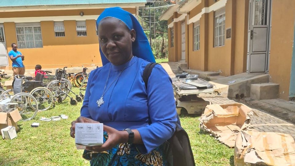 Eine Ordensschwester in Uganda zeigt Medikamente aus einer Hilfslieferung; im Hintergrund stehen Rollstühle.