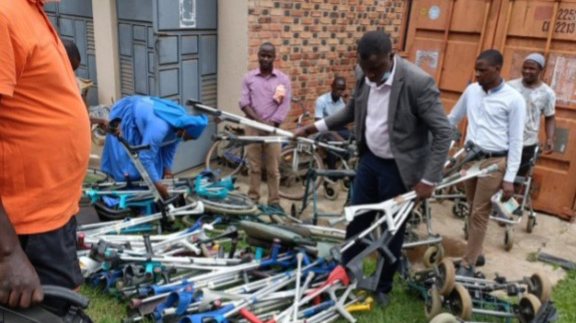 Helfer in Uganda sortieren neben einem Überseecontainer einen großen Stapel Gehhilfen und Krücken.